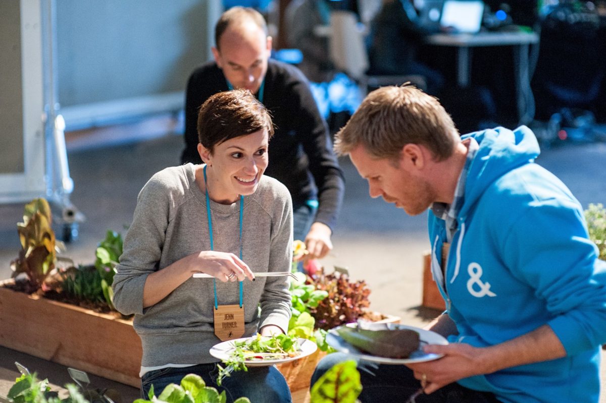 Me at a developer event wearing my name badge, grinning while I pick fresh greens from a raised garden bed with tongs, next to an attendee in a blue hoodie